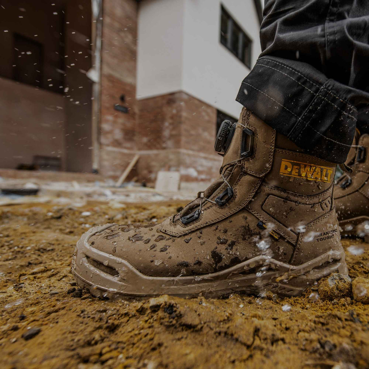 A close-up of a muddy DEWALT work boot being worn, with water splashing around it, set against an outdoor construction background.