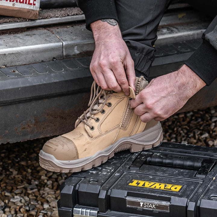 A person wearing a tan work boot with black pants, sitting on a truck bed next to a black DeWalt storage case.