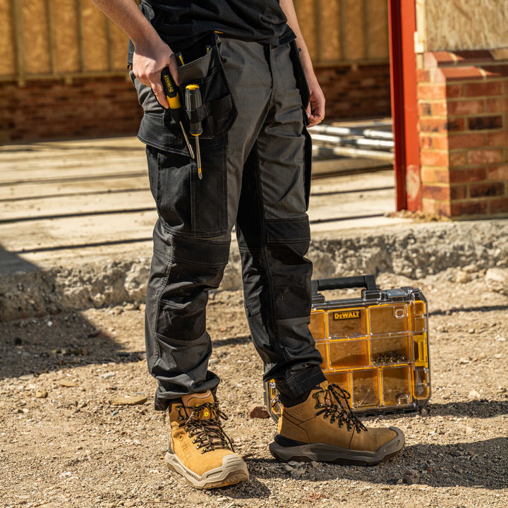 Person wearing work pants and boots with a tool belt and toolbox on a construction site.