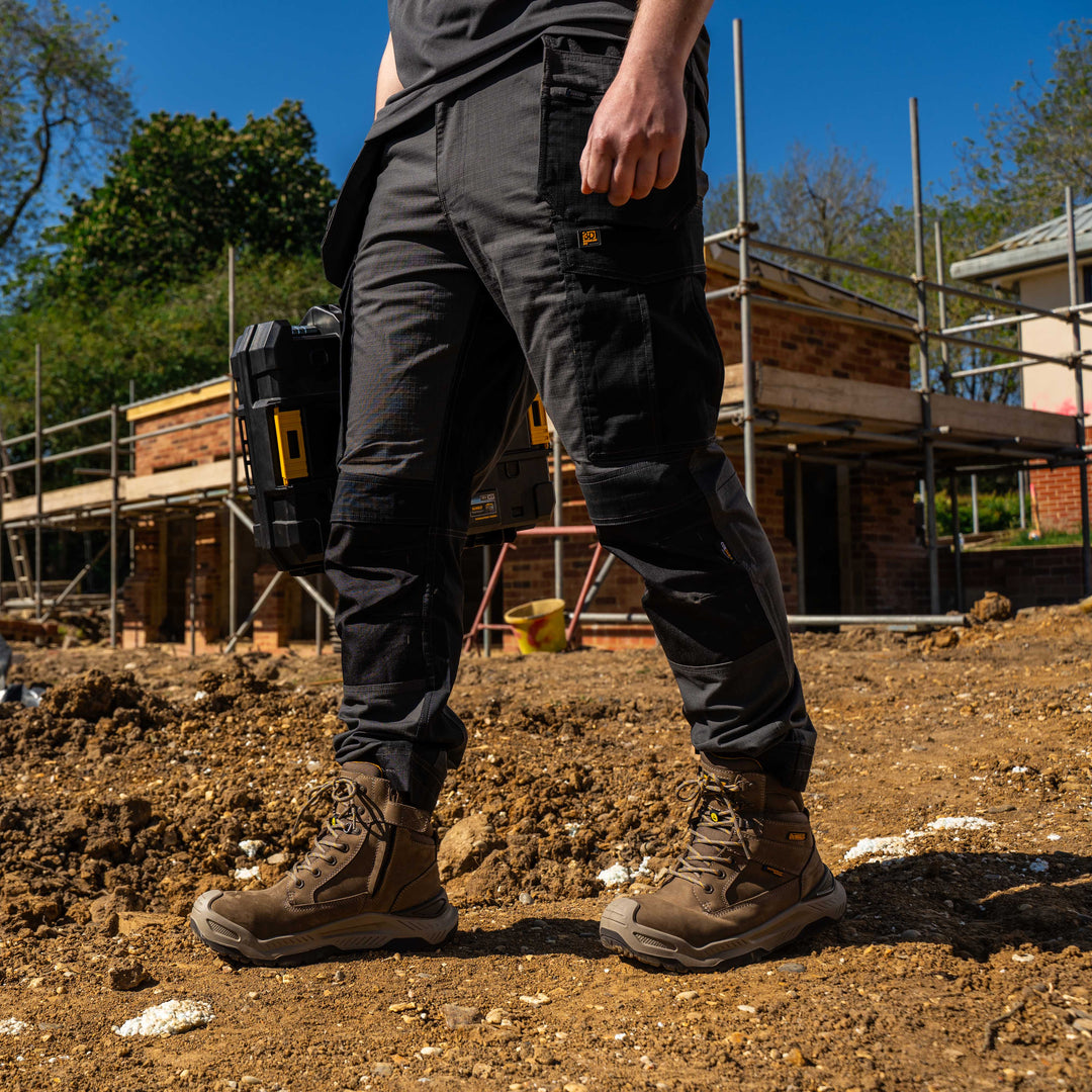 Person wearing black pants and brown boots on a construction site with scaffolding and building materials in the background.
