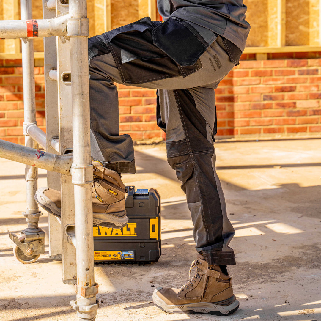 Person on a construction site wearing DeWalt boots and tools, with a brick wall in the background.