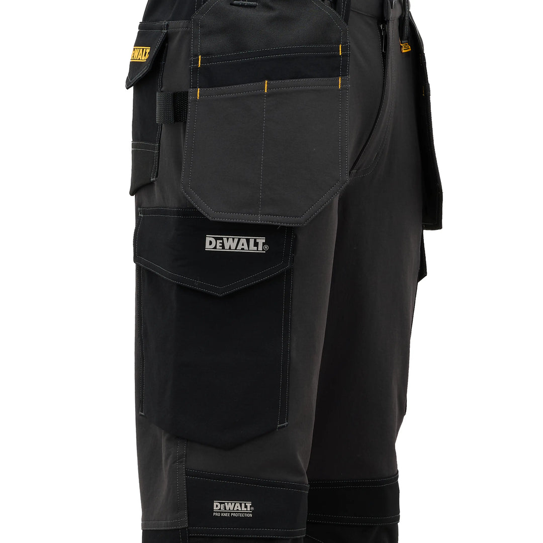 Close-up of black DeWalt work pants showing multiple utility pockets, yellow contrast stitching, and the DeWalt logo on the front pocket.