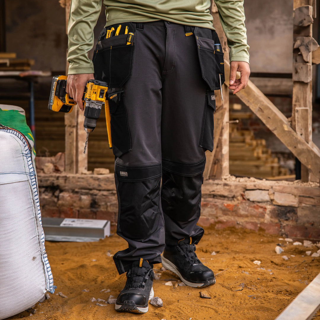 A person wearing dark gray and black work pants with multiple utility pockets and knee reinforcement stands on a construction site, holding a yellow cordless drill.