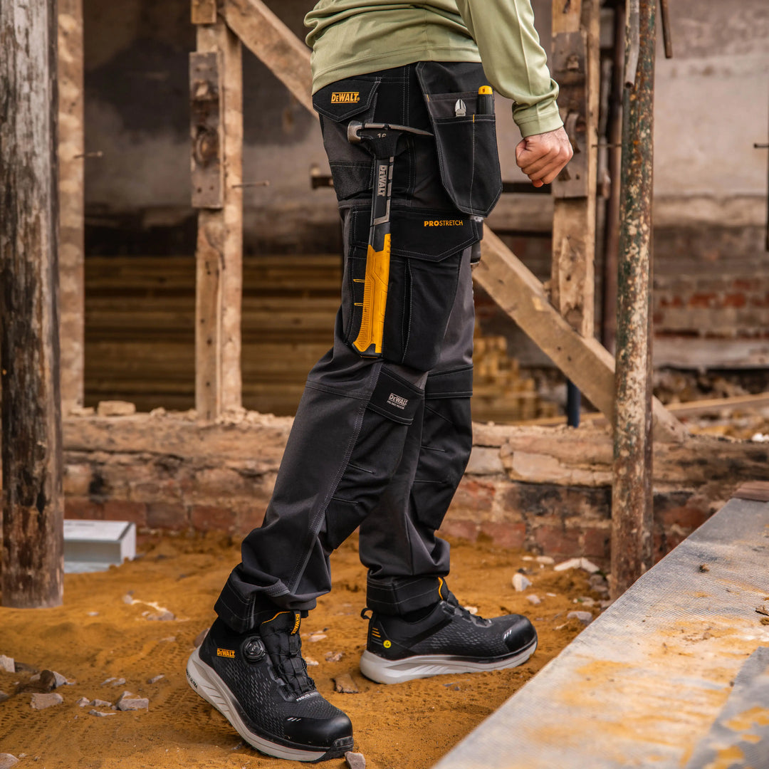 A person wearing dark gray and black DeWalt work pants with yellow accents, a matching tool belt, and black work boots, standing in a dusty construction site environment.