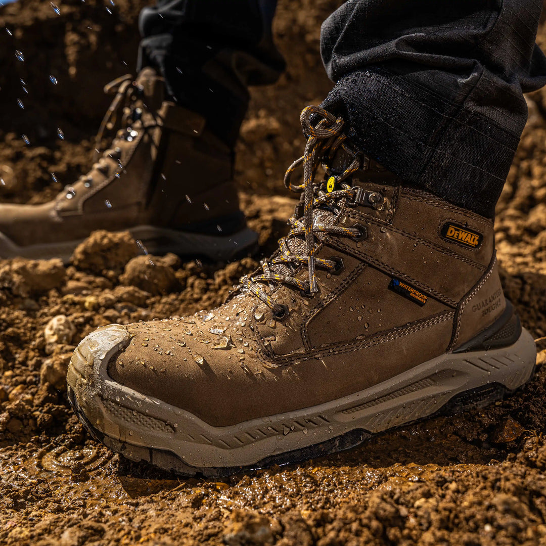 A close-up of a muddy brown DeWalt work boot being worn in wet dirt, with water droplets splashing off the boot. The DeWalt logo is visible on the side of the boot.