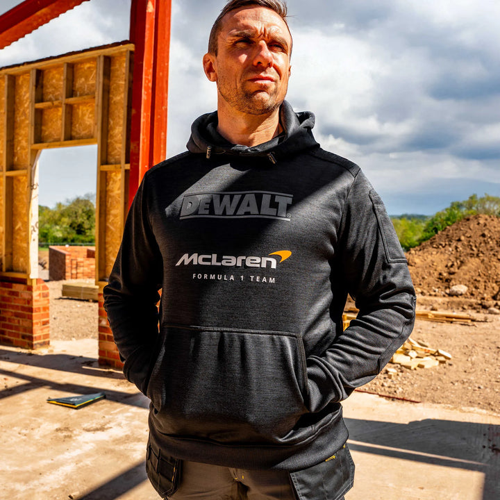 A man wearing a black DEWALT McLaren Formula 1 Team pullover hoodie stands outdoors in front of a construction site under a cloudy sky.