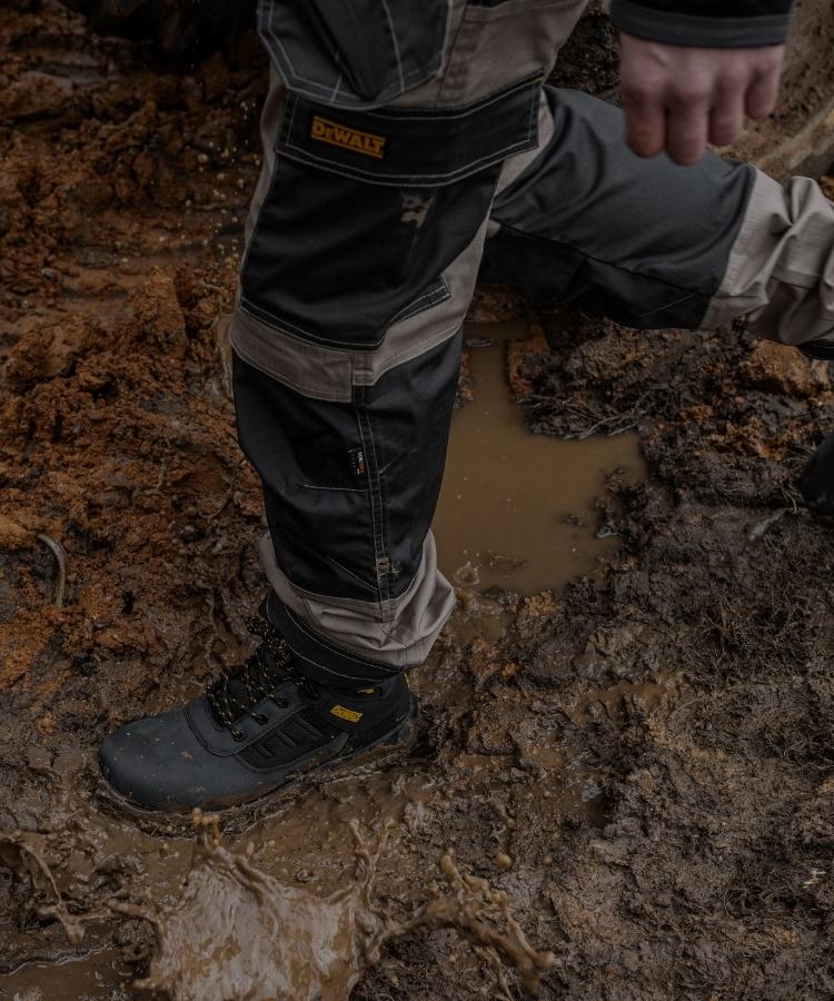 A close-up of a person wearing heavy-duty work pants and boots while walking through muddy, wet terrain. The pants are dark gray and black with a visible DeWalt logo patch on the upper thigh.