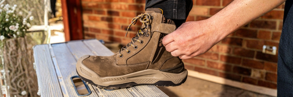 Person putting on a brown work boot with a brick wall and toolbox in the background